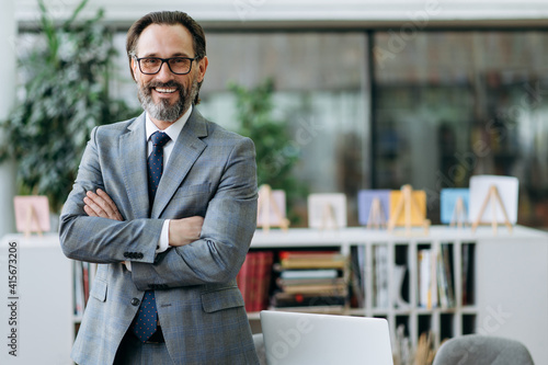 Portrait of a confident middle aged entrepreneur in stylish suit and eyeglasses standing in modern office, looking at the camera and friendly smiling