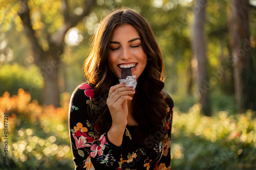 Gorgeous woman eating bar of dark chocolate in the sunny park. Woman enjoying chocolate with her eyes closed 