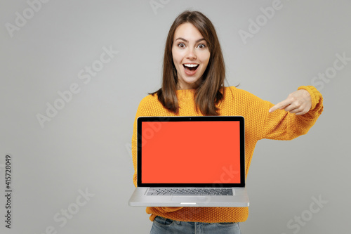 Young freelancer copywriter cute attractive woman 20s in knitted yellow sweater point index finger on laptop pc computer with blank screen workspace area isolated on grey background studio portrait..