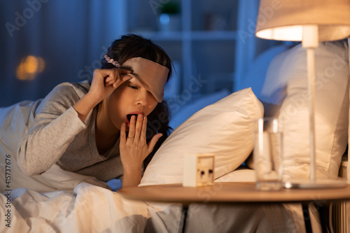 people, bedtime and rest concept - sleepy asian woman with clock yawning in bed at night