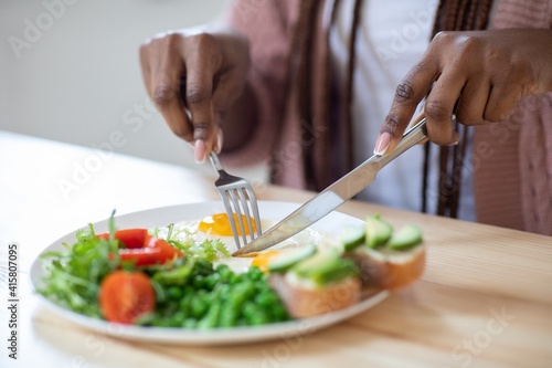 Unrecognizable african woman eating tasty breakfast in kitchen, using fork and knife