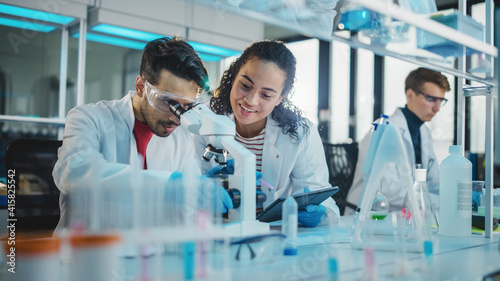 Modern Medical Research Laboratory: Portrait of Latin and Black Young Scientists Using Microscope, Digital Tablet, Doing Sample Analysis, Talking. Diverse Team of Specialists work in Advanced Lab