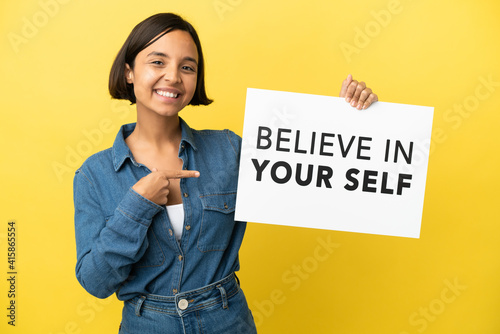 Young mixed race woman isolated on yellow background holding a placard with text Believe In Your Self and pointing it