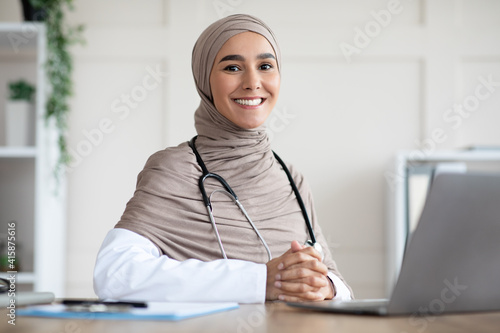 Cheerful doctor in hijab posing at her workdesk in clinic