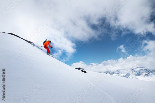 Young male skier in an orange suit rushes at speed along a snowy slope against the backdrop of high snow-capped mountains
