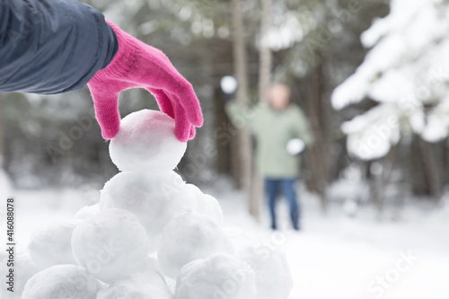 Original winter photograph of a woman's pink gloved hand picking up a snowball from a snowball pile for to have a snowball fight with man in the distance.