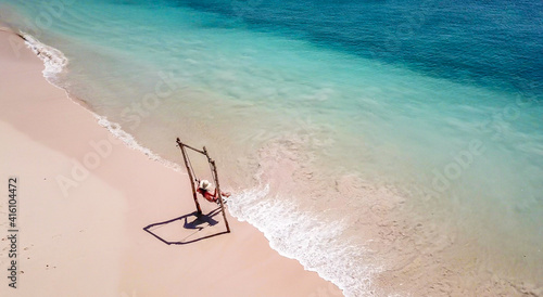 A girl swinging on a swing placed on the seashore of Pink Beach, Lombok, Indonesia. The swing has simple wood construction. Waves wash the pillars of it. In the back there are few boats. Drone capture