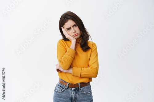 Sad and gloomy young woman sulking and frowning, holding hand over face and feeling upset, disappointed or offended, standing against white background