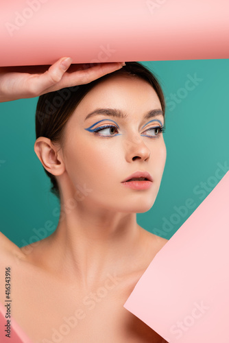 young woman with creative makeup looking away near hole in pink paper isolated on green