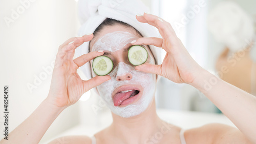 Cheerful woman with a towel on her hair and in a clay face mask and cucumbers on her eyes. Taking care of beauty at home