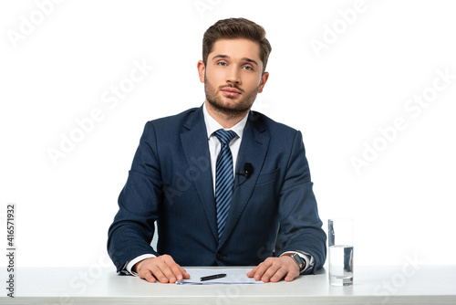 news anchor looking at camera while sitting at workplace near papers and glass of water isolated on white