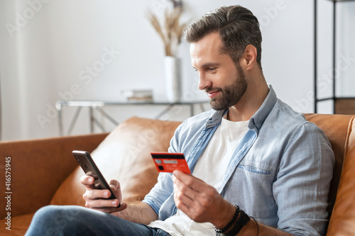 Bearded hipster guy businessman, holding mobile phone and credit card for online shopping and entering number to make payment, paying for utilities, using a virtual cabinet to manage banking account