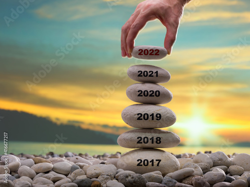 Happy new year 2022 concept. Years written on the rising stone pile. Man hand adding stone to tower. Background is blurred sunset sky