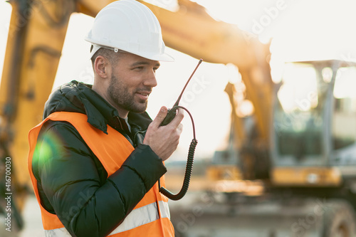 Young engineer talking radio communication (walkie talkie) and wearing a white helmet and construction orange vest. Close up engineers working on a building site with the sunny background.