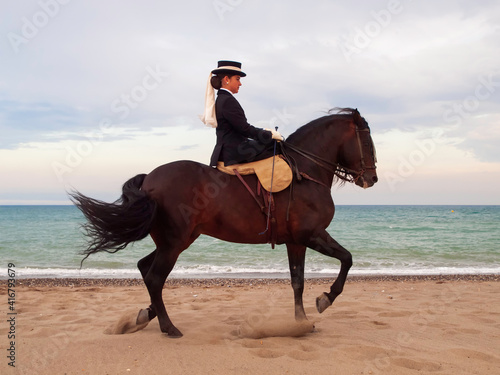 Joven jockey vestida de doma clasica montando un caballo marron en la playa