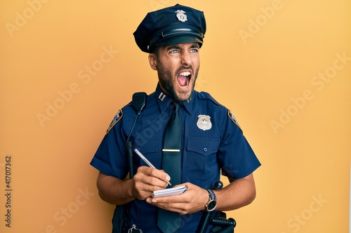 Handsome hispanic man wearing police uniform writing traffic fine angry and mad screaming frustrated and furious, shouting with anger. rage and aggressive concept.