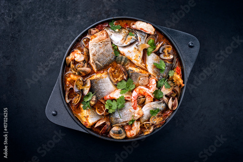 Modern style traditional Spanish seafood zarzuela de pescado with fish, king prawns and venus clams served in clear sauce as top view in design pot with copy space