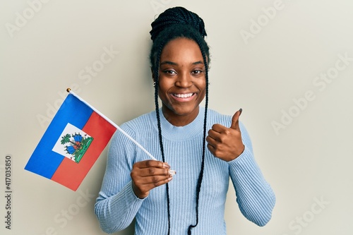 African american woman with braided hair holding haiti flag smiling happy and positive, thumb up doing excellent and approval sign