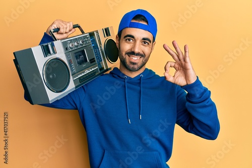 Young hispanic man holding boombox, listening to music doing ok sign with fingers, smiling friendly gesturing excellent symbol