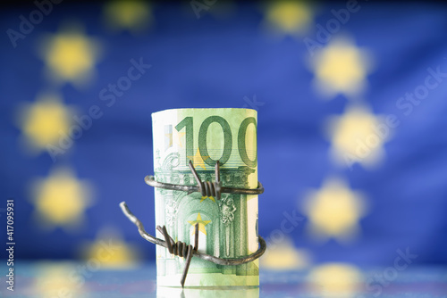 European Union currency wrapped in barbed wire against flag of EU as symbol of Economic warfare, sanctions and embargo busting.