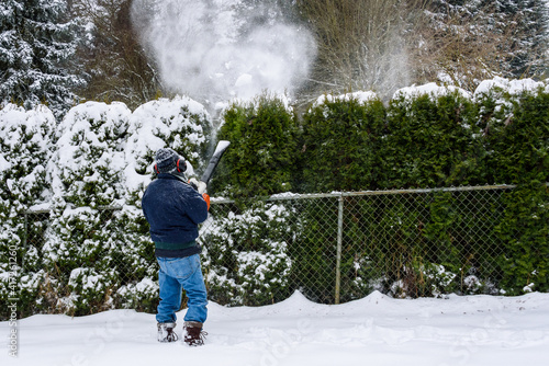 Snowy day, senior man using a snow-blower to remove snow from an arborvitae hedge 