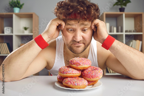 Funny athlete tempted by delicious doughnuts forgets about sports workout. Hungry man who loves sweet unhealthy junk foods sitting at table and looking at tempting fatty donuts with sprinkles on plate