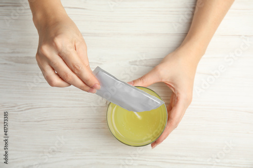 Woman pouring powder from medicine sachet into glass with water at table, top view