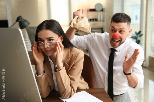 Man popping paper bag behind his colleague in office. Funny joke