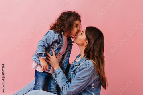 Brunette young mom kissing child. Studio shot of preteen girl with dark hair expressing love to mother.