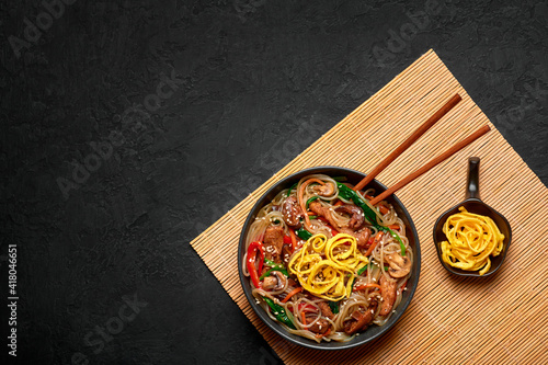 Japchae in black bowl on dark slate table top. Korean cuisine glass chapchae noodles dish with vegetables and meat. Asian traditional food. Authentic meal. Top view. Copy space