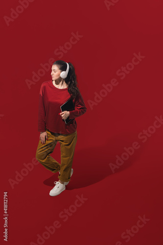 Dancing with headphones. High angle view of young woman on red studio background. Human emotions and facial expressions concept. Full length portait, copyspace for ad. Fashion, retro style.