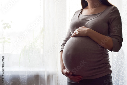 Pregnant woman with big belly at window. Maternity prenatal care and woman pregnancy concept. Young pregnant woman holds her hands on her swollen belly. Love concept. Horizontal, copy space.