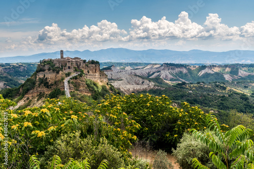 Civita 01 - borgo antico su sperone di tufo con fiori gialli, calanchi montagne e nuvole