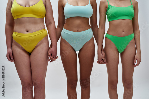Cropped shot of three women with different body shapes in colorful underwear holding hands, supporting each other, standing together isolated over light background