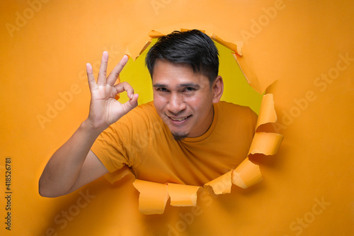 Young entertaining man through torn yellow paper hole showing okay sign with facetious of content and happiness.