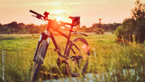 The mountain bike stands on a gravel bike path among green vegetation illuminated by the rays of the setting sun.