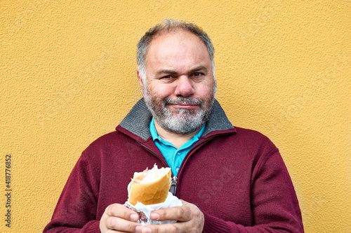 Funny bearded mature man eating sandwich on the street against a yellow wall. Man hungry like a wolf biting bread. Unhealthy eating.