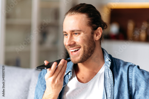 Speakerphone conversation, audio message recording. Handsome young caucasian bearded guy holding a mobile phone in his hand, talking on a speakerphone with a girl or friend, smiling, sits at home