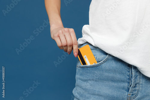 Close up cropped photo portrait shot of caucasian female hand arm putting credit bank card in jeans pants denim pocket isolated on dark blue background studio portrait. Money finance currency concept.