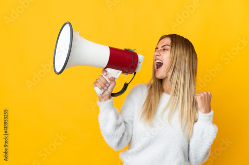 Young blonde woman isolated on yellow background shouting through a megaphone to announce something in lateral position