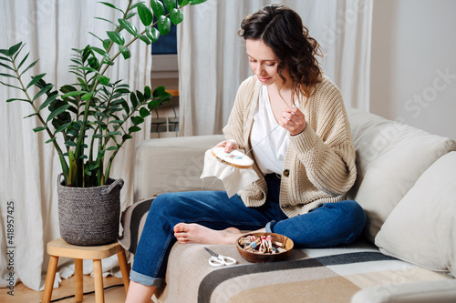 Happy young woman sewing in comforts of her apartment, embroiders a picture