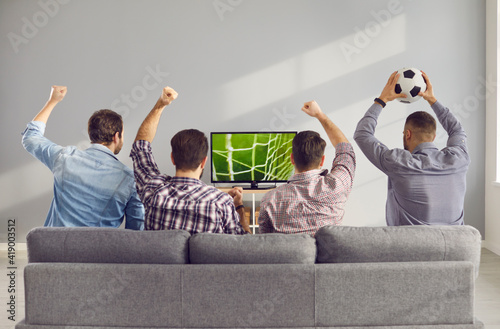 Four adult male friends with arms raised emotionally watching a football match on TV at home. Unrecognizable friends sit on the couch with their backs to the camera and cheer for their favorite team.