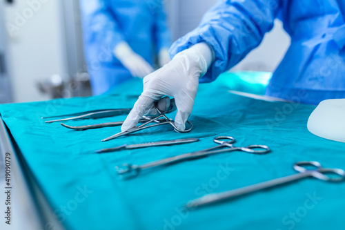 Nurse hand taking surgical instrument for group of surgeons at background operating patient in surgical theatre. Steel medical instruments ready to be used. Surgery and emergency concept