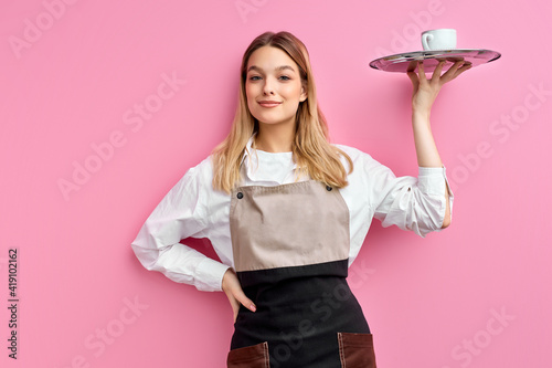 nice woman waitress in apron, offering cup of delicious tasty coffee on tray, stand smiling, friendly staff of restaurant. isolated over pink studio background