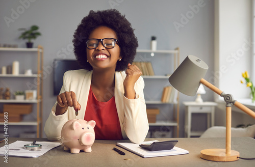 Money saving for shopping concept. Lovely satisfied, excited happy young african american woman in eyewear with yes gesture putting coin into piggybank because of income exceeds expenses significantly