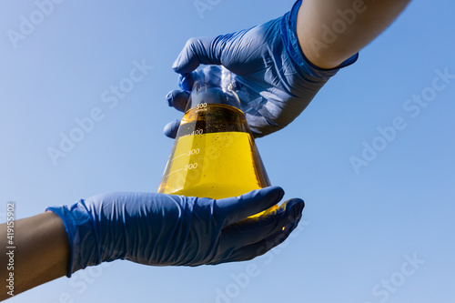 Hand with gloves holding beaker with ethanol biofuel against blue sky