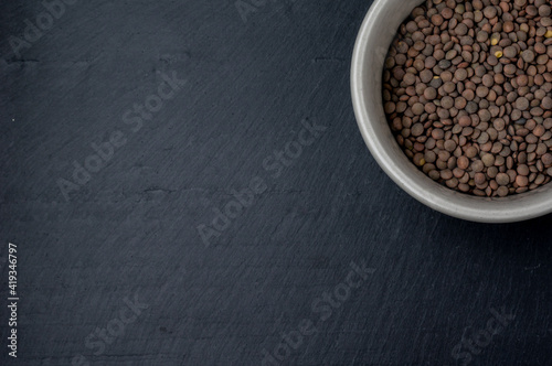 top view of a fragment of a white ceramic bowl filled with fresh lentils on the top right edge of a dark slate surface that is the background of the frame and serves as a copy space.