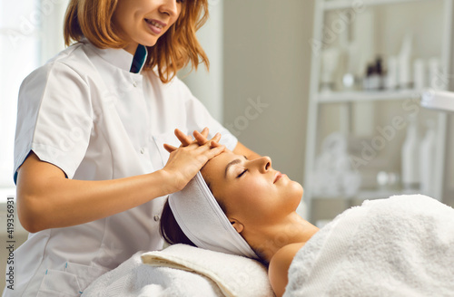 Cosmetologist or dermatologist making facial beauty massage treatment for young woman in beauty spa salon with fingers