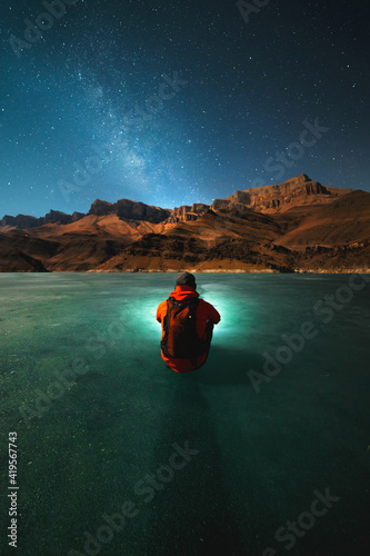 A young man with a backpack sits on the ice of a frozen lake at night in winter and looks at the mountains and the milky way by the light of the moon