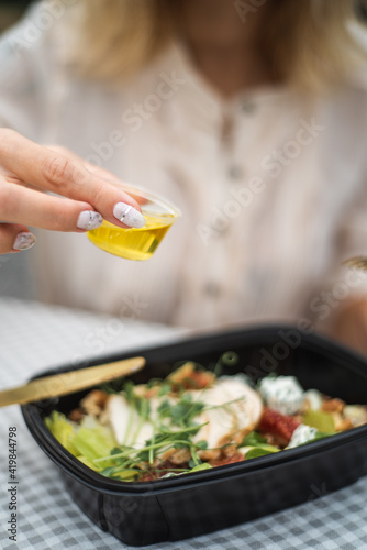 Woman hand pouring flavored olive oil to fresh vegetable salad. Preparation healthy vegetarian food for garden party outdoors. The salad is in a food container. Food delivery.